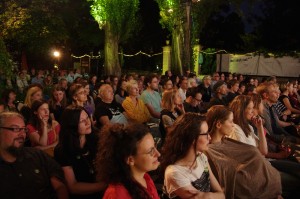 Zuschauerinnen und Zuschauer einer Filmvorführung auf dem dotdotdot Open Air Kurzfilmfestival in Wien. Im Hintergrund bunte Lichter und Bäume bei Nacht.