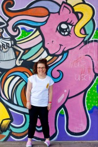 Ninia Binias steht vor einer Graffiti-Wand. Im Hintergrund ein pinkes Einhorn mit bunten Haaren aufgemalt. Ninia trägt ein weißes T-shirt, schwarze Hose, pinke Schuhe und rote Brille.