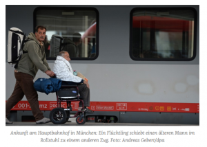 Ein Mann schiebt einen Mann im Rollstuhl. Hinter ihnen ein Zug der Deutschen Bahn. Beide Männer sind Flüchtlinge und befinden sich am Münchner Hauptbahnhof.