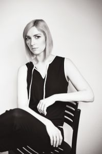 MArlies Hübner sitzt auf einem Stuhl. Sie hat blonde kurze Haare und dunkle Augen, sie trägt einen schwarzen Jumpsiut. Es ist ein Bild in schwarz-weiß.