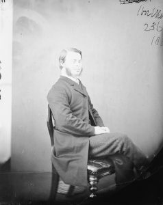 Eine Schwarz-Weiß-Fotografie aus dem 19. Jahrhundert. Ein junger Mann mit Down-Syndrom sitzt in chiquer Kleidung, also Stoffhose und Frack, auf einem Holzstuhl. Er trägt einen langen Bart auf beiden Seiten der Wange und hat die Haare im Seitenscheitel nach hinten gekämmt.