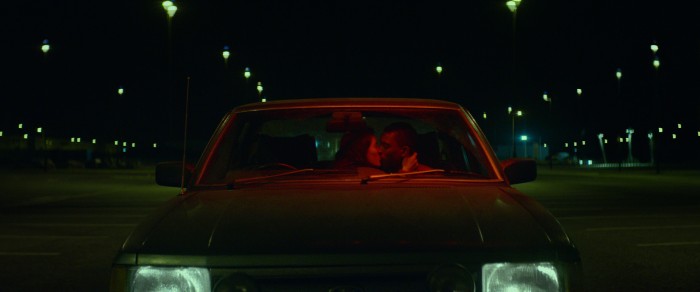 Ein küssendes Pärchen sitzt im Auto, welches auf auf einem leeren und beleuchteten Parkplatz steht.