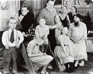 Behinderte Menschen aus dem Film "Freaks" 1931. In der Mitte steht ein nicht behinderter Mann mit Schnurrbart. Um ihn herum behinderte Menschen, meist kleinwüchsig. Eine Schwarzweiß-Fotografie.