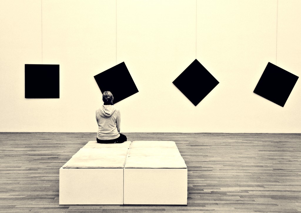 Frau sitzt im Museum, man blickt auf ihren Rücken. Sie sitzt auf weißen Polstermöbeln. Sie schaut auf 4 schwarze Quadrate, wovon 3 schief hängen und eins gerade.