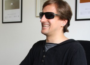 Foto: Heiko Kunert mit schwarzer Sonnenbrille. Er hat braune kurze Haare und trägt einen silbernen Ohrring. Er ist von der Seite aufgenommen und lächelt. Im Hintergrund Bilder in einem Büro.