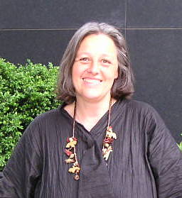 Foto: Heike Oldenburg. Schulterlange schwarz-graue Haare, eine schwarze Bluse, eine bunte Kette und ein Lächeln im Gesicht. Im Hintergrund grüner Busch.