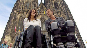 Eine Rollstuhlfahrerin und ein Rollstuhlfahrer stehen vor dem Kölner Dom. Sie unterhalten sich.