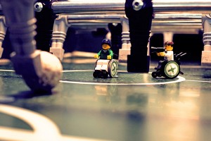 Zwei Legofiguren im Rollstuhl auf einem Kicker-Fußballtisch fotografiert, er von beiden hält eine Videokamera in der Hand