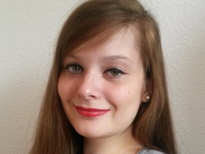 Porträtfoto von Judyta Smykowski. Sie hat lange braune Haare, grüne Augen, Sommersprossen und roten Lippenstift. Vor einer weißen Wand lächelt sie in die Kamera.