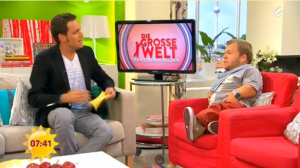 Screenshot aus dem Sat.1-Frückstücksfernsehen. Der Moderator interviewt Michel Arriens zur Serie "Die grosse Welt der kleinen Menschen". Alles ist sehr bunt: die Tapete neongrün und weiß, der rote Sessel, auf dem Michel sitzt, ist knallrot.