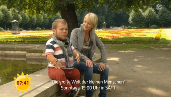 Screenshot aus der Anmoderation der Serie "Die große Welt der kleinen Menschen" mit Michel Arriens und Ulla Kock am Brink. Beide sitzen nebeneinander auf dem Gemäuer eines Springbrunnes in einem Park voller Blumen, die Sonne scheint.