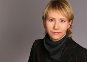 Profilfoto von Karin Chladek. Sie hat schulterlange blonde Haare, trägt einen grauen Rollkragenpullover unter einem schwarz-rot gestreiften Jacket und lächelt in die Kamera.