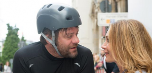Georg ("Der Kotzbrocken") trägt einen Helm und ist auf seinem Handbike, schaut Sophie tief in die Augen, sie lächelt.