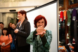 Apothekerin Simonitsch zeigt auf einer Veranstaltung lächelnd die Gebärde für "Apotheke". Dafür hält sie ihre rechte Faust mit erhobenem Daumen auf ihrer linken Handfläche. Sie hat rote kinnlange Haare und trägt ein grünes Oberteil mit Blumen.