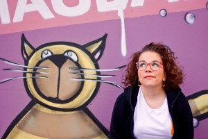 Ninia Binias sitz mit offenen lockigen Haaren, roter Brille, weißem T-shirt und schwarzer Jacke vor einer Graffiti-Wand. Sie lächelt ähnlich wie die Katze, die hinter ihr auf die Wand gemalt wurde. Der Hintergrund der Wand ist pink und lila.