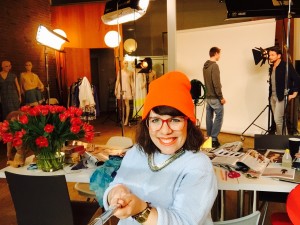 Ninia Binias als Moderatorin im Studio von RTL für Aufnahmen zum Fashionmag. Sie trägt eine rote Mütze, rote Brille, helleblauen Pullover und lacht in die Kamera. Im Hintergrund arbeiten Kameraleute am Set.