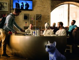 Drei Männer sitzten an einem großen Tisch, in der Mitte der Rapper Cro mit Pandamaske. Sie werden von einem Kellner bedient. Etwas abseits vom Tisch steht eine weiße Hundefigur (Mops). Der Hund trägt eine Brille