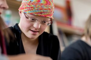 Eine Frau mit Down-Syndrom schaut freundlich nach unten. Sie trägt eine Brille, ein buntes Kopftuch und ein schwarzes Oberteil.