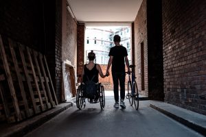 Ein Pärchen geht Hand in Hand durch einen Tunnel. Die Frau sitzt im Rollstuhl und der Mann schiebt ein Fahrrad.