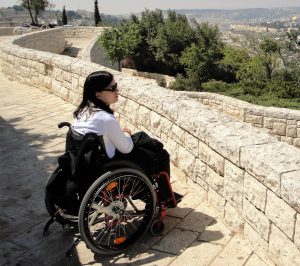 Laura Chrobok hat schwarze Haare, trägt ein weißes Shirt und eine schwarze Hose. Sie sitzt im Rollstuhl und schaut über eine Mauer hinweg in die Ferne.