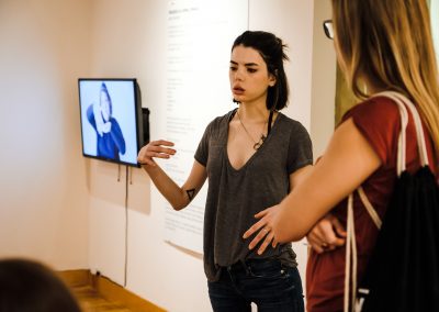 Eine Frau mit kurzen schwarzen Haaren ist zu sehen. Sie erklärt einer anderen Frau mit langen blonden Haaren die Ausstellung.