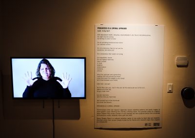 An der Wand ist ein Gedicht, daneben ein Bildschirm mit einer Frau die das Schriftliche gebärdet.