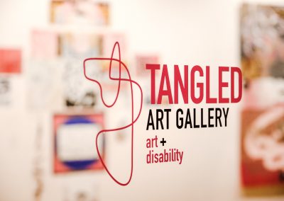Zu sehen ist das Logo der Tangled Art Gallery. Es besteht aus pinken Linien die verbinden sind. Außerdem der Schriftzug Tangled Art Gallery art+disability.