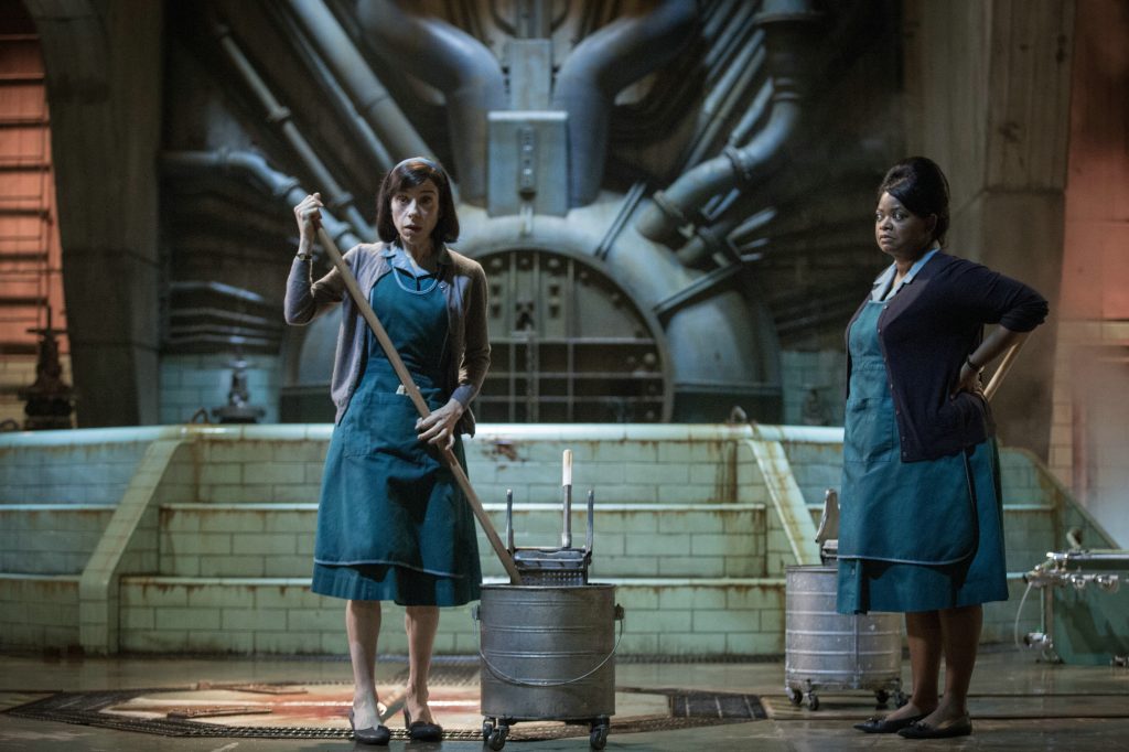 Screenshot aus dem Film Shape of Waters. Zwei Frauen stehen in einer Forschungseinrichtung nebeneinander und schauen verängstigt. Die Frau links im Bild hält einen Wischmob in den Händen und taucht diesen in den Putzeimer.