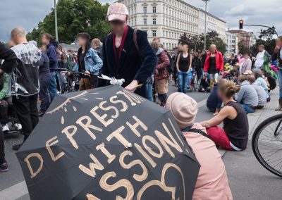 Zu sehen sind etliche Demonstrierende auf der Pride Parade. Zwei Teilnehmer*innen sitzen auf dem Gehweg unter einem Regenschirm, der mit dem Motto "Despression with Passion" verziert ist.