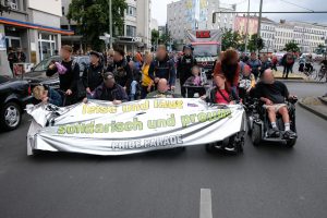 Mehrere Rollstuhlfahrer*innen, die ein Transparent mit dem Titel "leise und laut - solidarisch und proud!"