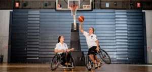 Anne Patzwald und Mareike Miller in der Sporthalle, in der die WM ausgetragen wird. Sie sitzen beide im Rollstuhl, unter dem Basketballkorb. Mareike Miller steht dabei nur auf zwei Rädern und ist seitlich angekippt und so versucht sie den Ball in den Korb zu legen.