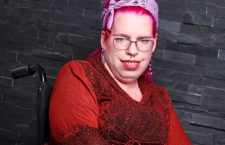 Tanja sitzt im Rollstuhl, trägt ein rotes Kleid und ein rosanes Haarband, hat rosane Haare und eine Brille.