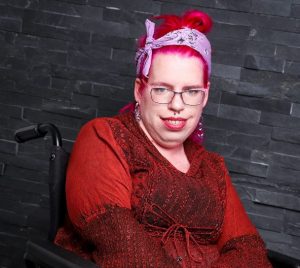 Tanja sitzt im Rollstuhl, trägt ein rotes Kleid und ein rosanes Haarband, hat rosane Haare und eine Brille.