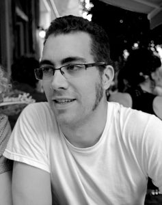 Ein schwarz-weiß Bild von Mikael Ross. Er hat kurze schwarze Haare, trägt eine Brille und ein weißes T-Shirt