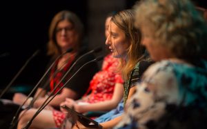 Jovana Komnenic nimmt bei einer Podiumsdiskussion in Alice Springs teil. Jovana hat schulterlanges blondes Haar und spricht gerade in Mikrophone.Sie sitzt zwischen vier anderen Menschen, die ebenfalls Mikrophone vor sich stehen haben. Die anderen sind nur in der unschärfe zu erkennen.
