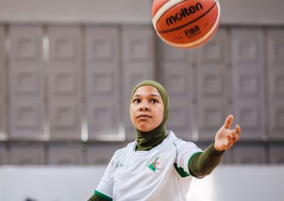 Eine Rollstuhlbasketballspielerin trägt ein olive-farbenes Kopftuch und ein weißes t-Shirt. Sie wirft einen Basketball in die Luft.