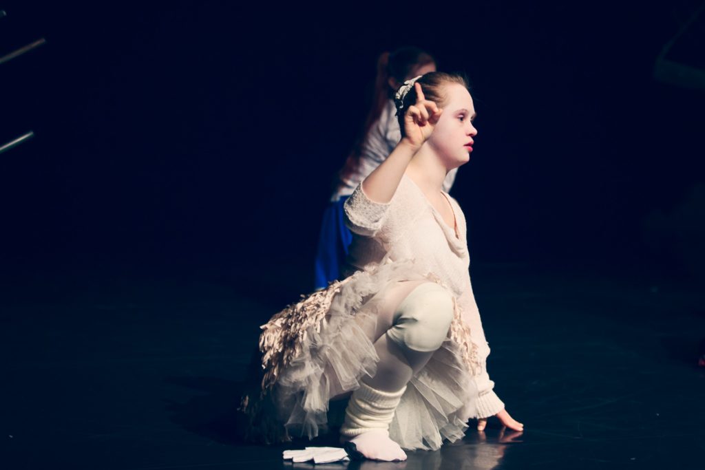 Die Tänzerin Nele Buchholz kniet in einem weißen Kleid auf der Bühne und hebt den Zeigefinger.