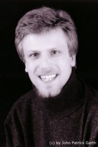 Schwarz-Weiß Foto von John Patrick Garth. Er trägt eine dunkle Jacke und hat kurzes, dubkelblondes Haar.