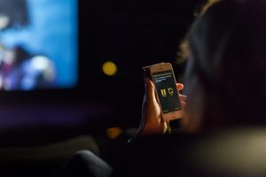 Eine Person sitzt im Kino und hat das Smartphone mit der Greta-App in der Hand. Es ist dunkel und im Hintergrund ist die Leinwand in der unschärfe zu sehen.