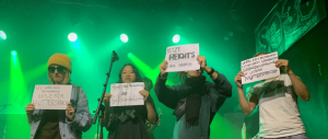 Auf einer Bühne stehen vier Aktivist*innen von Deaf Performance Now und halten Schilder mit ihren Forderungen hoch. Die Bühne ist grün angeleuchtet.