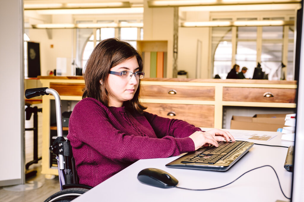 Eine Frau mit dunkelbraunen Haaren, Brille und dunkelrotem Pullover sitzt an einem Tisch am Computer. Sie sitzt im Rollstuhl.